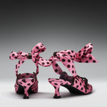 A pair of pink satin high-heeled sandals with black polka dots and large matching bow straps, shown from the back on a dark-to-light gray gradient studio background.