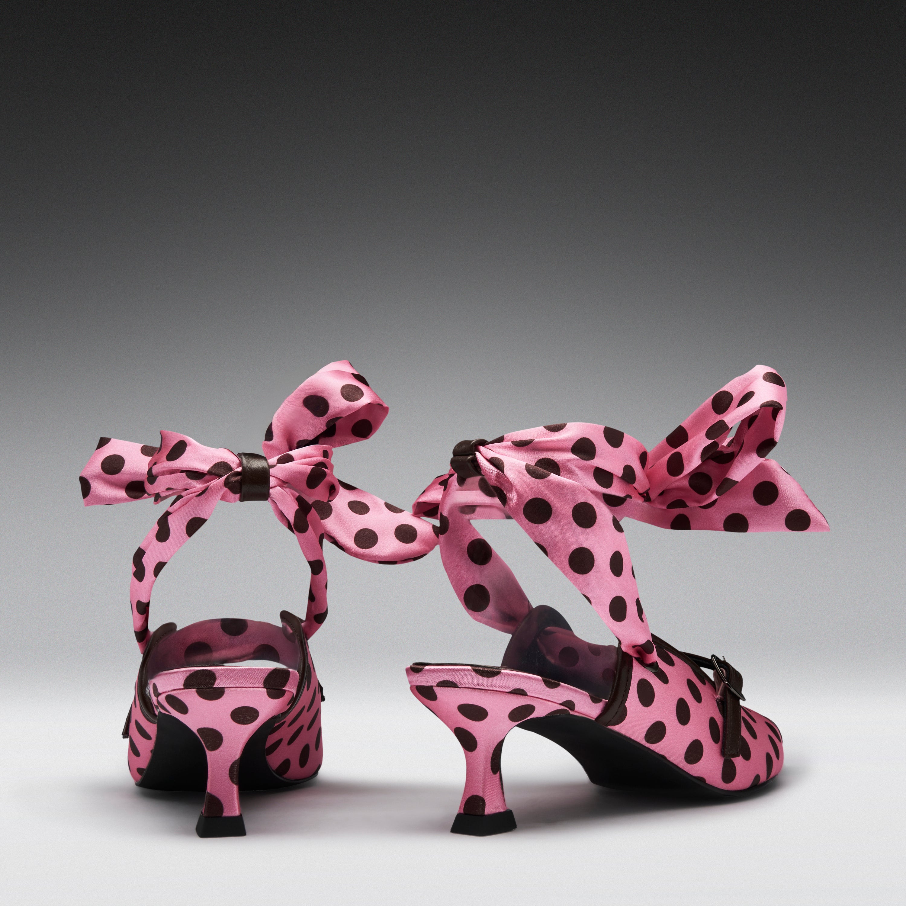 A pair of pink satin high-heeled sandals with black polka dots and large matching bow straps, shown from the back on a dark-to-light gray gradient studio background.