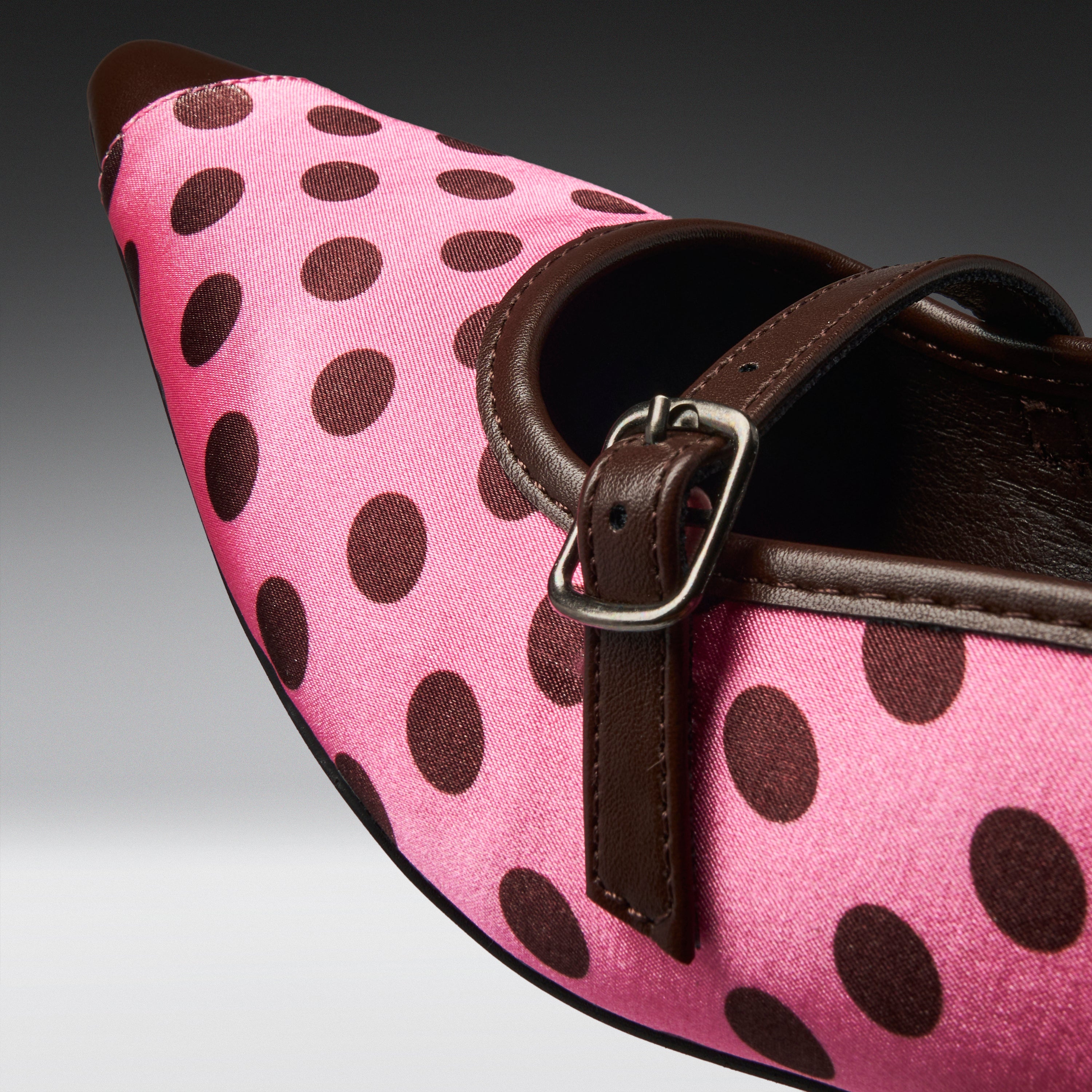 A close-up of a pink pointed-toe shoe with dark brown polka dots, a dark brown strap, and a silver buckle.