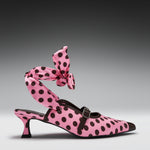 A pink pointed-toe heeled mule with black polka dots, featuring a matching polka-dot ankle strap tied in a large bow, photographed on a dark gray gradient studio background.