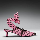 A pink pointed-toe heeled mule with black polka dots, featuring a matching polka-dot ankle strap tied in a large bow, photographed on a dark gray gradient studio background.