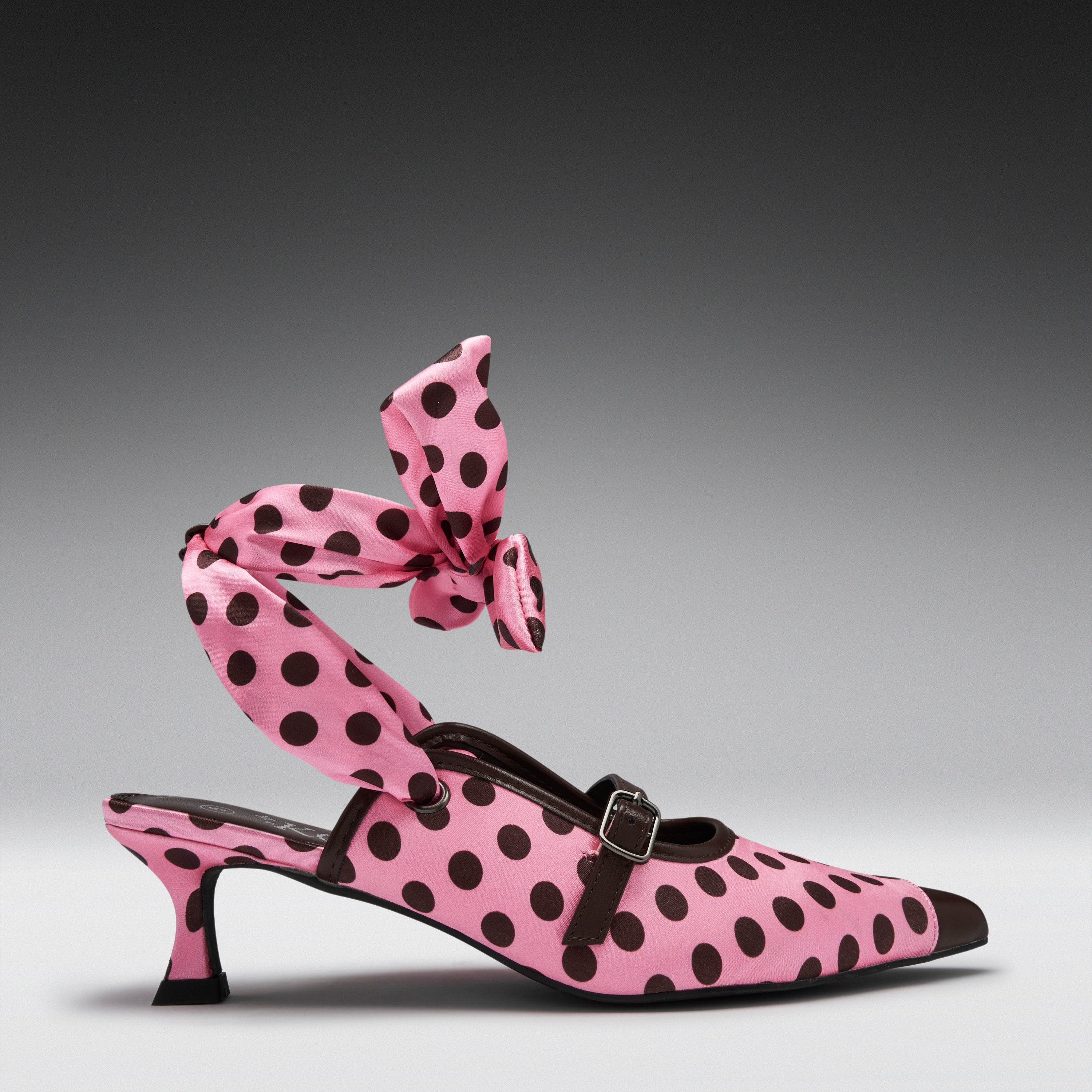 A pink pointed-toe heeled mule with black polka dots, featuring a matching polka-dot ankle strap tied in a large bow, photographed on a dark gray gradient studio background.
