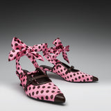 Pink pointed-toe Mary Jane-style heels with dark brown polka dots, bow ankle straps, and dark brown leather trim, photographed on a gray gradient studio background.