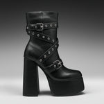 A black leather ankle boot with a chunky block heel, platform sole, and multiple decorative straps adorned with silver studs and buckles, shown against a dark gray gradient background.