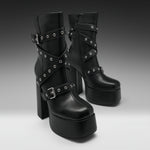 A pair of black platform ankle boots with high block heels, decorated with multiple crisscrossing straps and silver-tone metal grommets, shown against a dark-to-light gray gradient studio background.