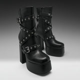 A pair of black platform ankle boots with high block heels, decorated with multiple crisscrossing straps and silver-tone metal grommets, shown against a dark-to-light gray gradient studio background.