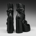 A pair of black platform ankle boots with chunky block heels, double platform soles, and multiple decorative straps with silver-tone grommets and buckles, shown from the back and side against a dark gray gradient background.
