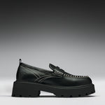 A single black leather loafer with a chunky lug sole, featuring contrast white stitching and a decorative strap across the vamp, shown against a dark gray gradient background.