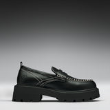 A single black leather loafer with a chunky lug sole, featuring contrast white stitching and a decorative strap across the vamp, shown against a dark gray gradient background.