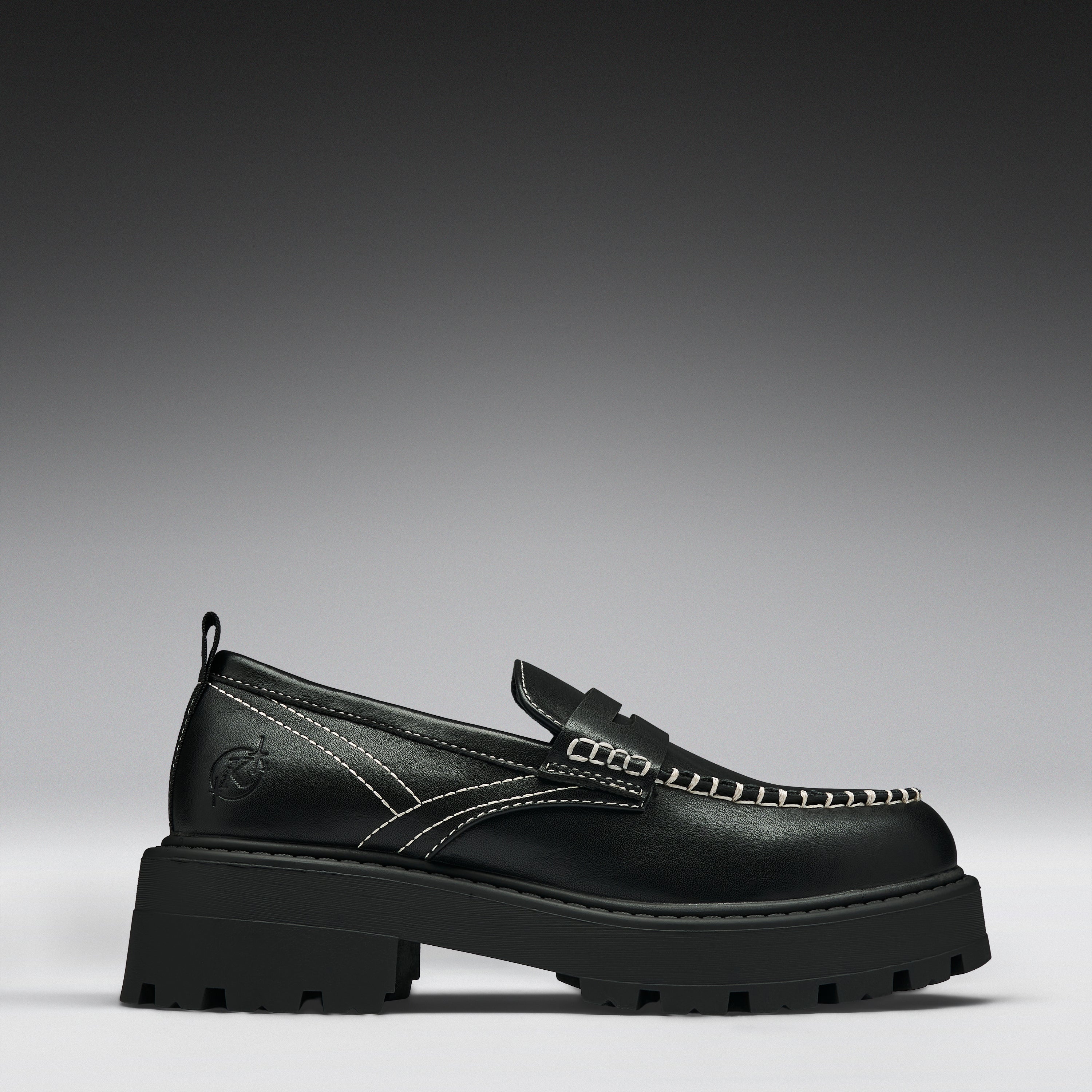 A single black leather loafer with a chunky lug sole, featuring contrast white stitching and a decorative strap across the vamp, shown against a dark gray gradient background.