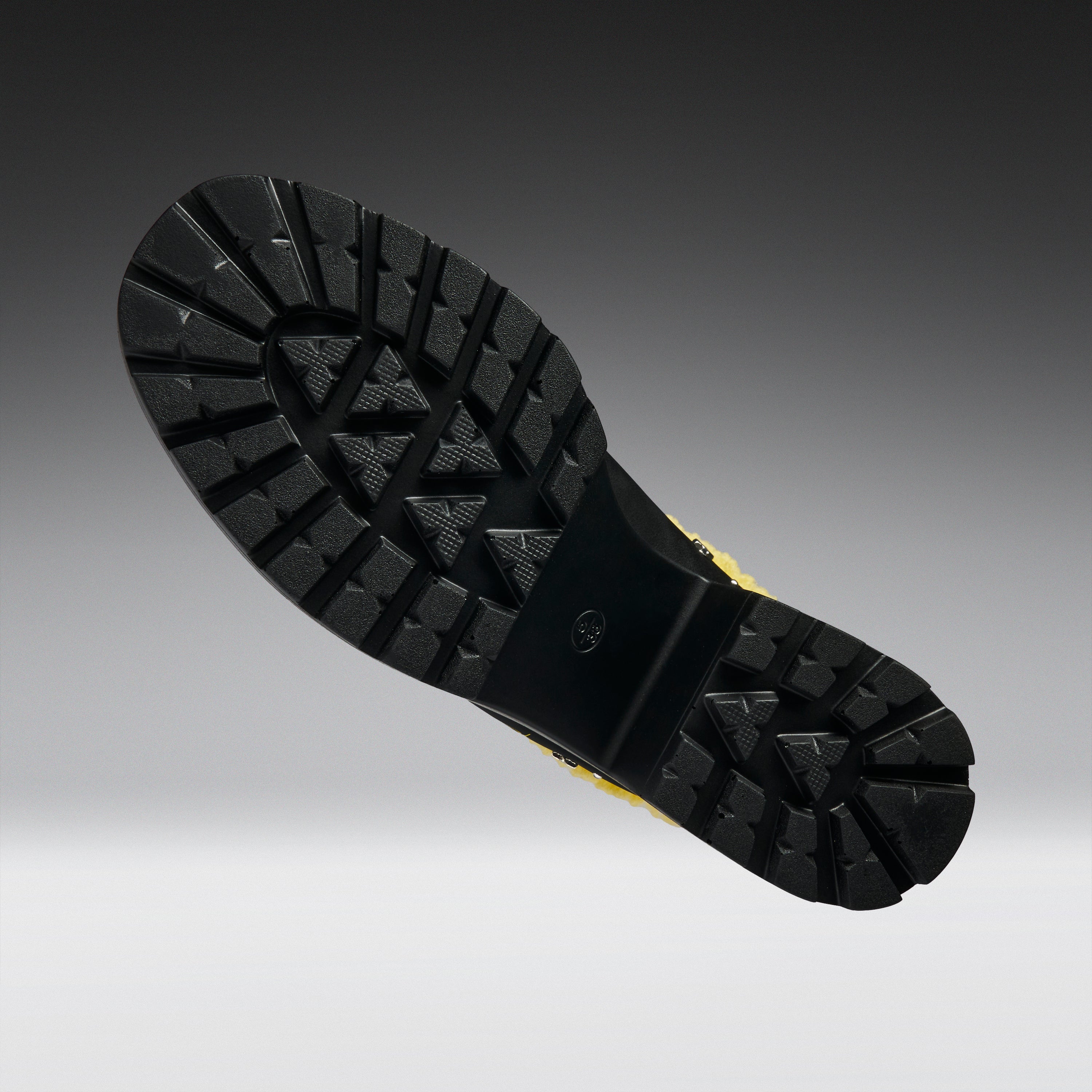 A close-up of a black rubber boot sole with deep tread patterns, shown against a dark gray to light gray gradient background.