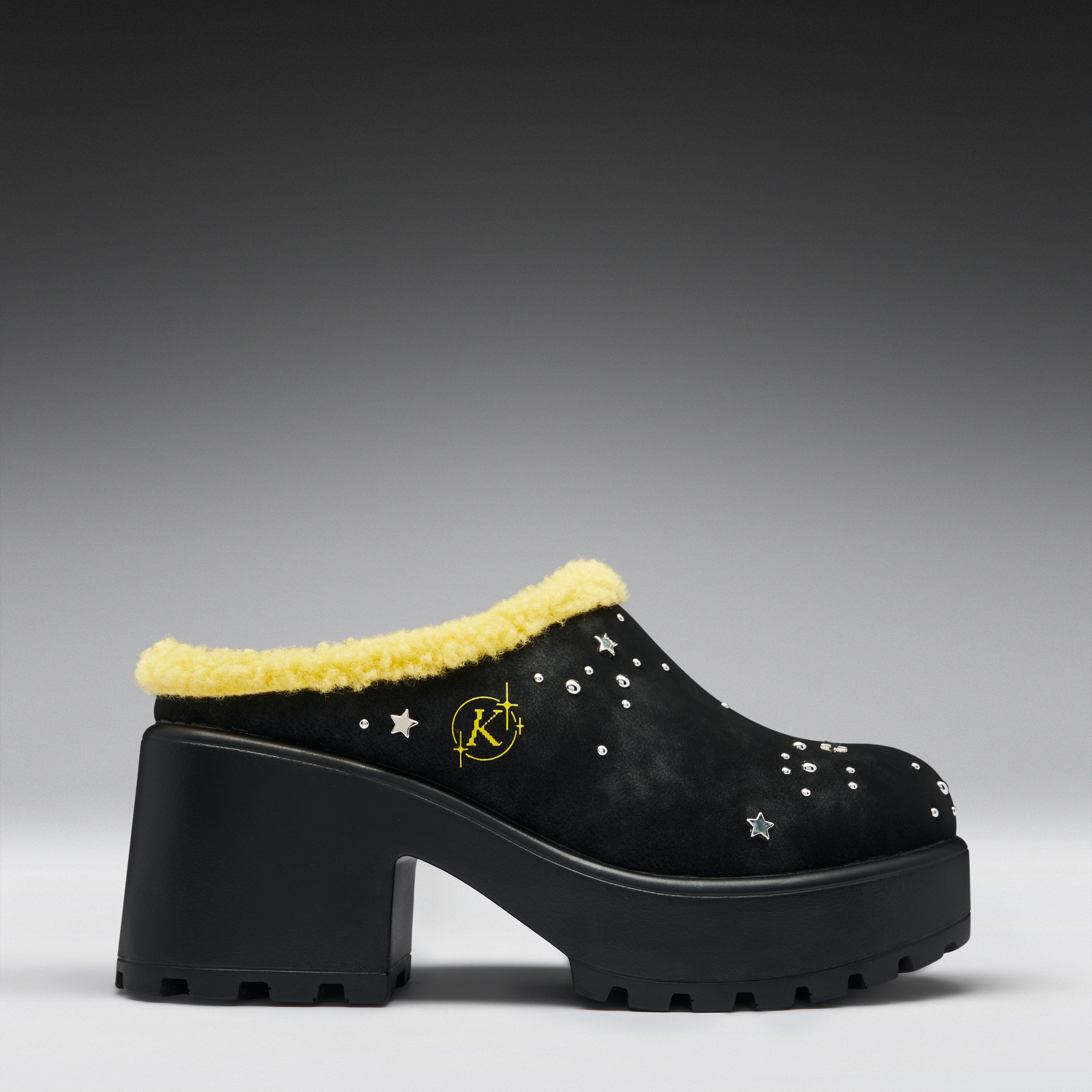 A black platform mule shoe with a yellow faux-fur trim, decorated with silver star-shaped studs and a yellow circular 'K' logo on the side, set against a dark gray gradient background.