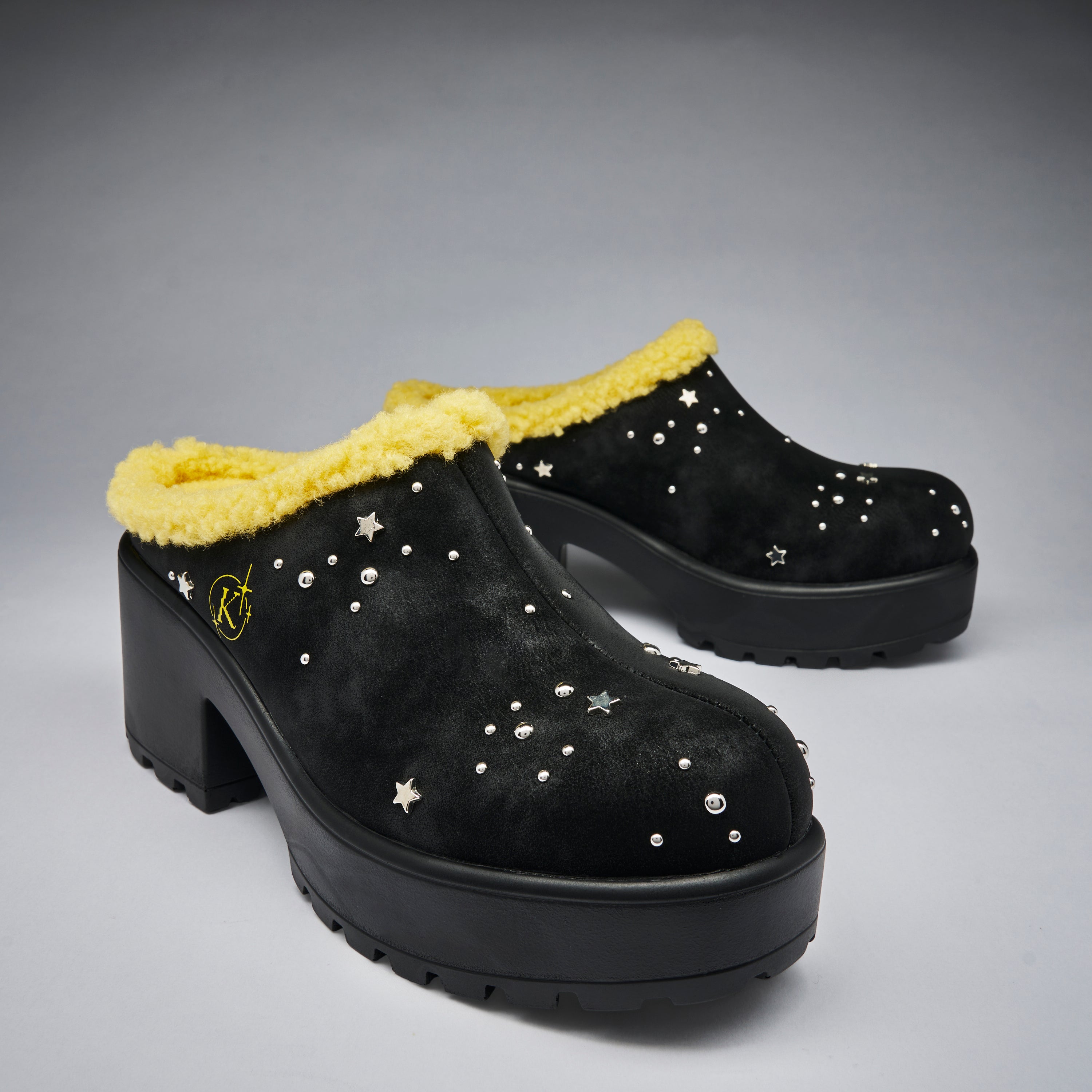 A pair of black platform mule slip-on shoes with a bright yellow faux-fur trim and silver star-shaped studs, displayed on a neutral gray background.