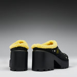 A pair of black platform mule shoes with bright yellow sherpa lining and small gold star embellishments, shown from the side against a dark gray gradient background.