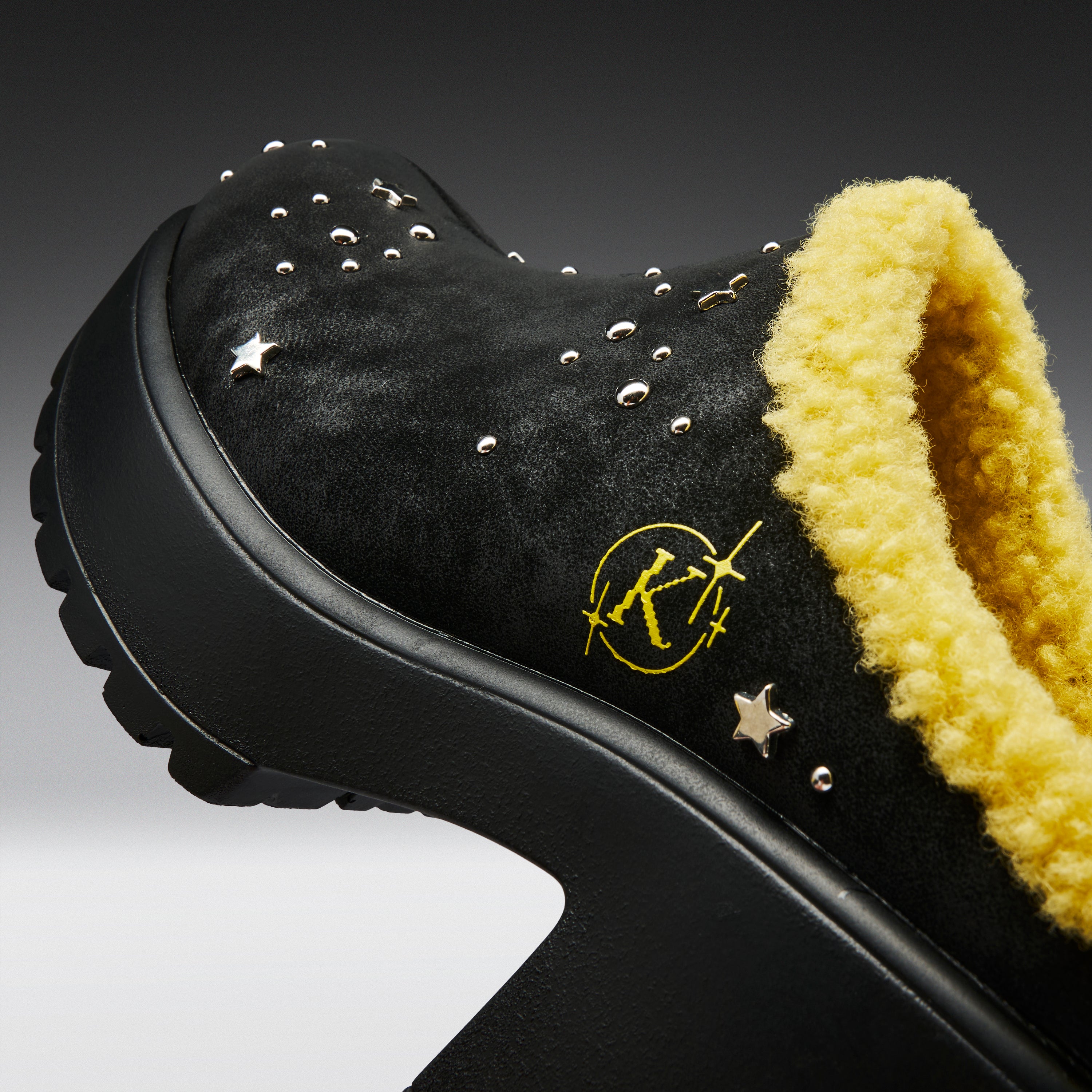 A close-up of a black platform mule shoe with a bright yellow sherpa lining, silver studs, and a yellow embroidered logo on the side.