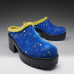 A pair of blue suede clogs with a yellow faux-fur trim, decorated with silver star and dot embellishments, shown on a gray gradient background.