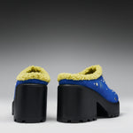 A pair of blue platform mule heels with thick black lug soles and pale yellow sherpa lining, shown from the back against a dark gray gradient background.