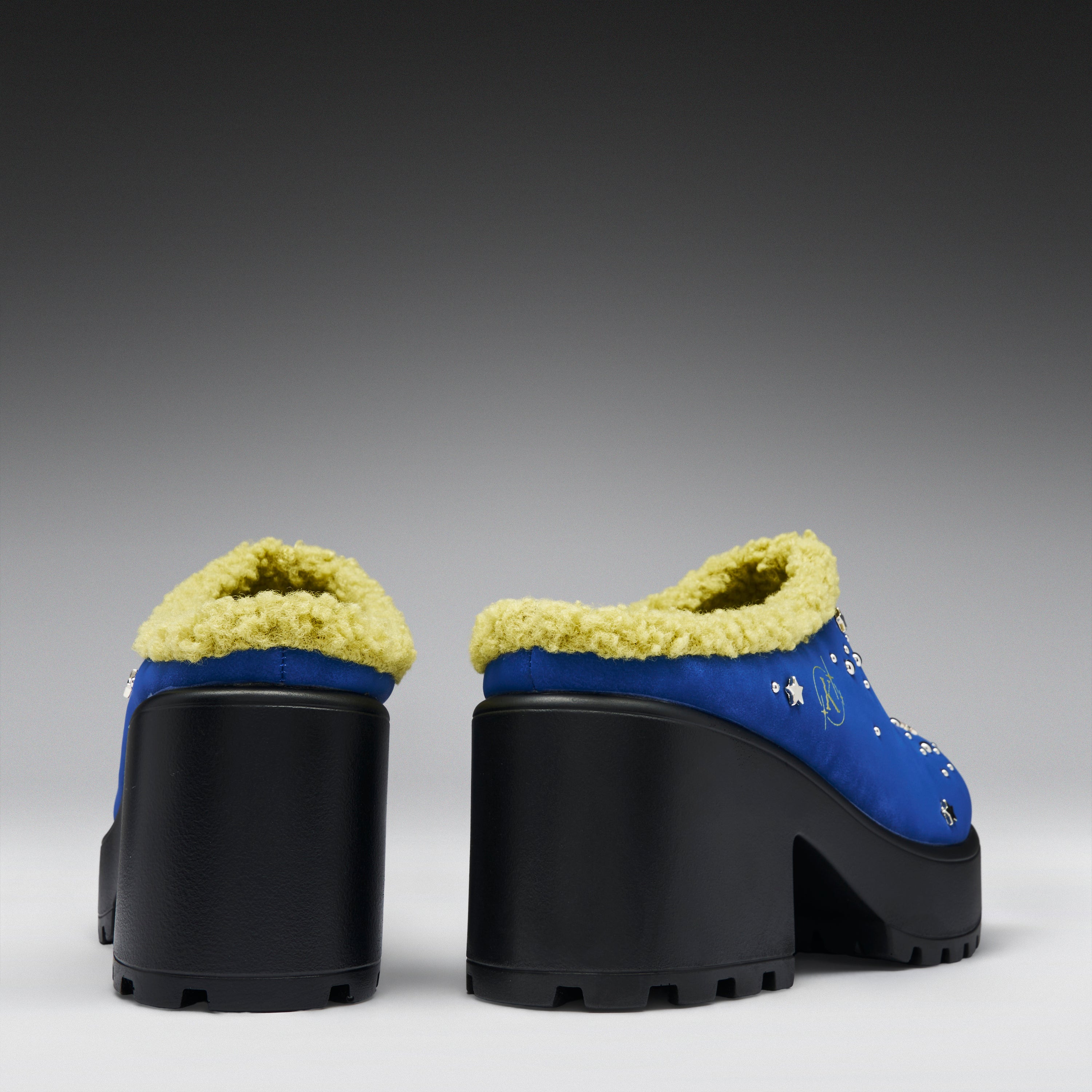 A pair of blue platform mule heels with thick black lug soles and pale yellow sherpa lining, shown from the back against a dark gray gradient background.