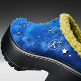 A close-up of a blue suede clog-style shoe with a black platform sole, a yellow-green sherpa lining, and silver star and stud embellishments.