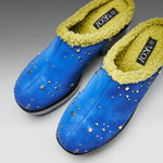 A pair of blue slip-on slippers with a yellow-green sherpa lining and silver star-shaped studs, shown on a neutral gray background.