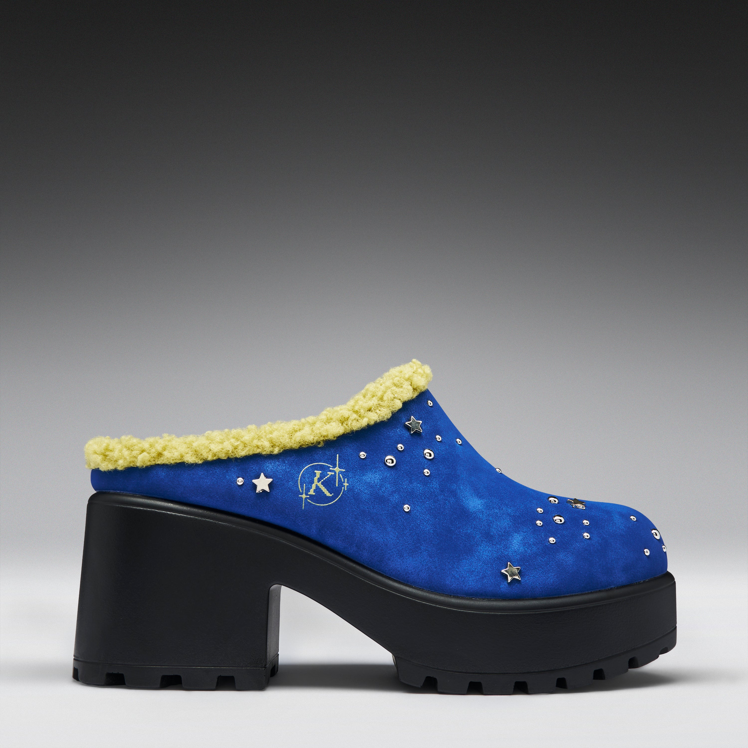 A blue suede mule clog with a chunky black platform sole, a yellow faux-fur trim, and silver star embellishments, shown against a dark gray gradient background.