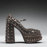 A single high-heeled platform Mary Jane shoe covered in metallic rose gold studs, shown in profile against a dark gray gradient background.