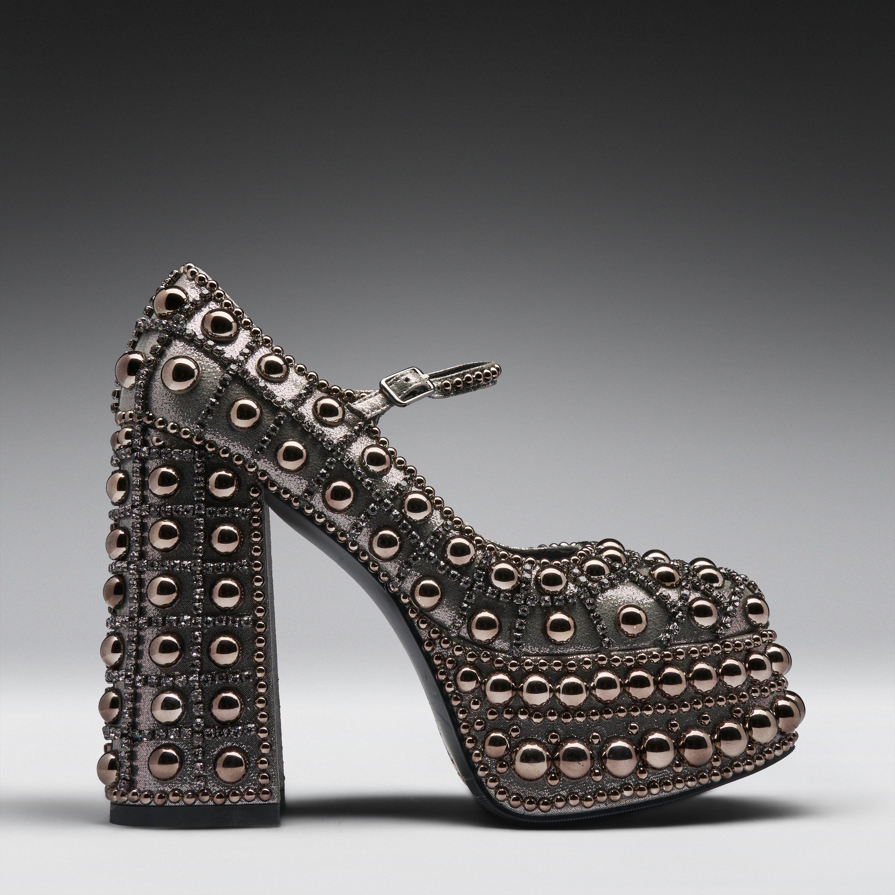 A single high-heeled platform Mary Jane shoe covered in metallic rose gold studs, shown in profile against a dark gray gradient background.