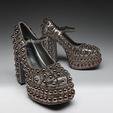 A pair of metallic silver platform Mary Jane shoes heavily embellished with rows of dark metallic studs and beads, set against a dark gray studio background.