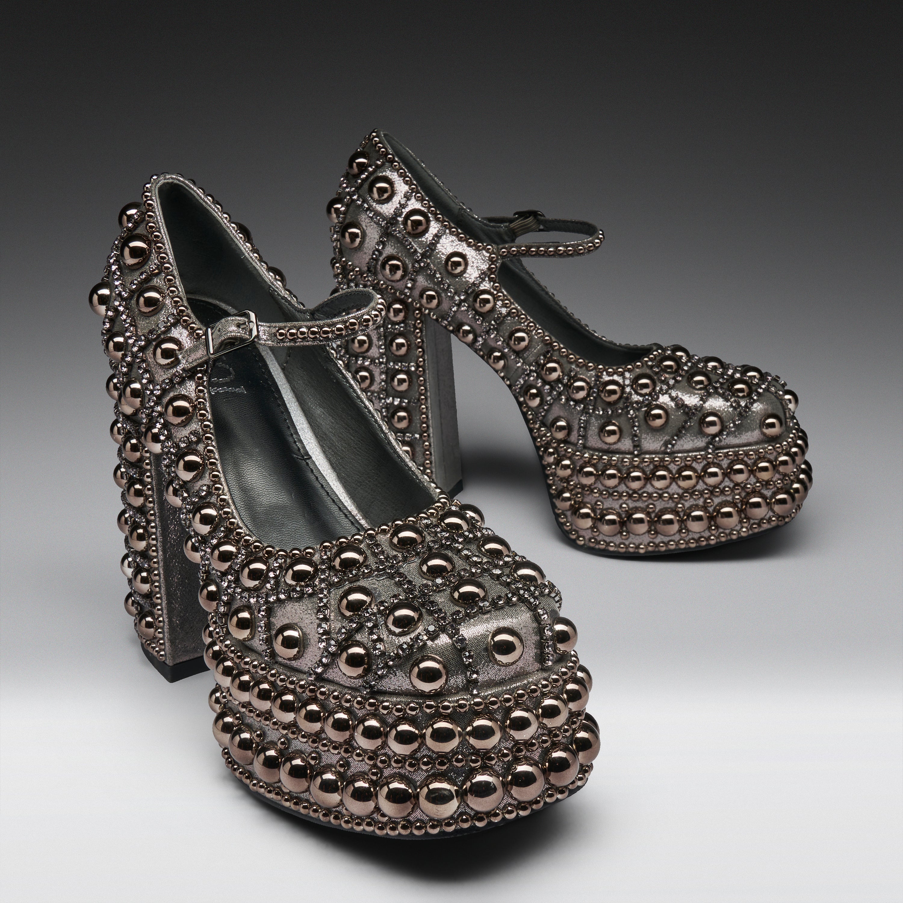 A pair of metallic silver platform Mary Jane shoes heavily embellished with rows of dark metallic studs and beads, set against a dark gray studio background.