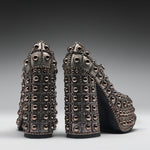 A pair of high-heeled platform shoes covered in metallic studs and rhinestones, shown from the back on a dark gray gradient studio background.