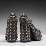 A pair of high-heeled platform shoes covered in metallic studs and rhinestones, shown from the back on a dark gray gradient studio background.