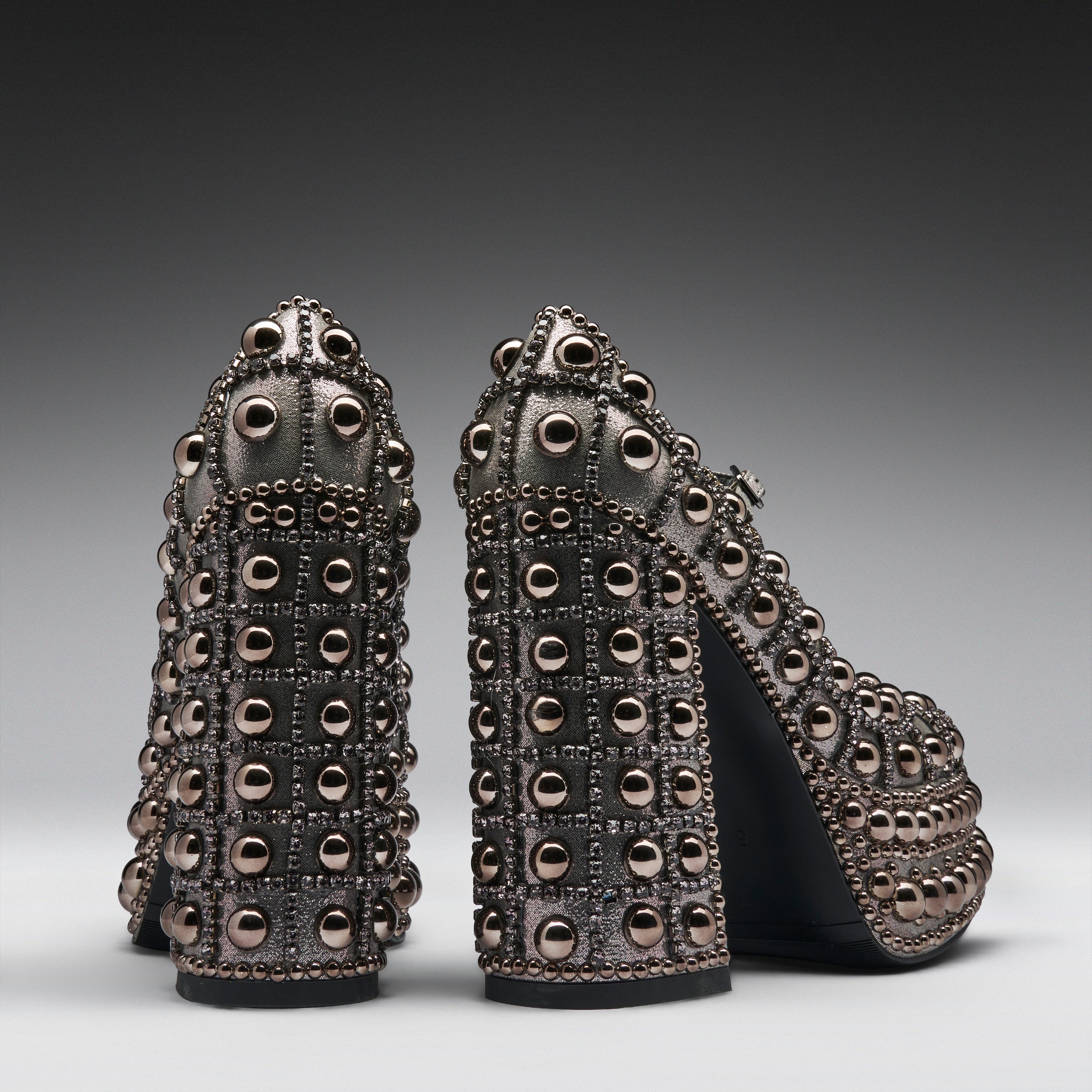 A pair of high-heeled platform shoes covered in metallic studs and rhinestones, shown from the back on a dark gray gradient studio background.