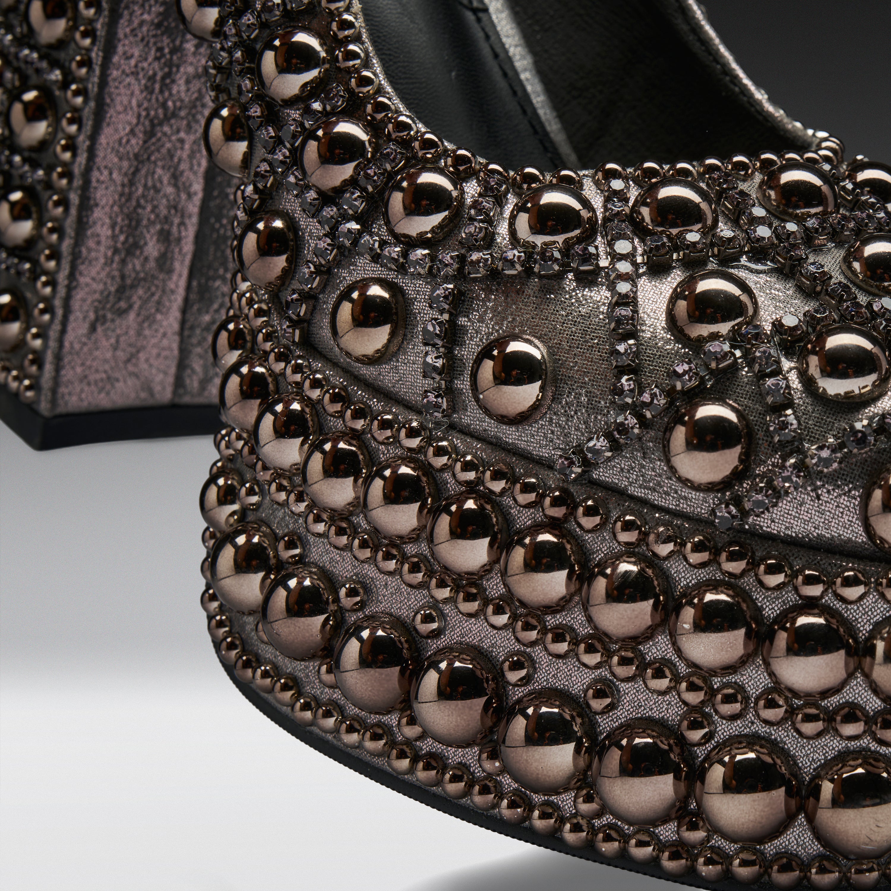 Close-up of a metallic studded shoe featuring rows of large rose gold dome studs and smaller dark metallic studs on a textured black and silver upper.