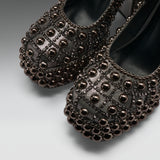 A pair of black leather ballet flats heavily embellished with metallic bronze and dark brown beaded studs, arranged on a light gray studio background.