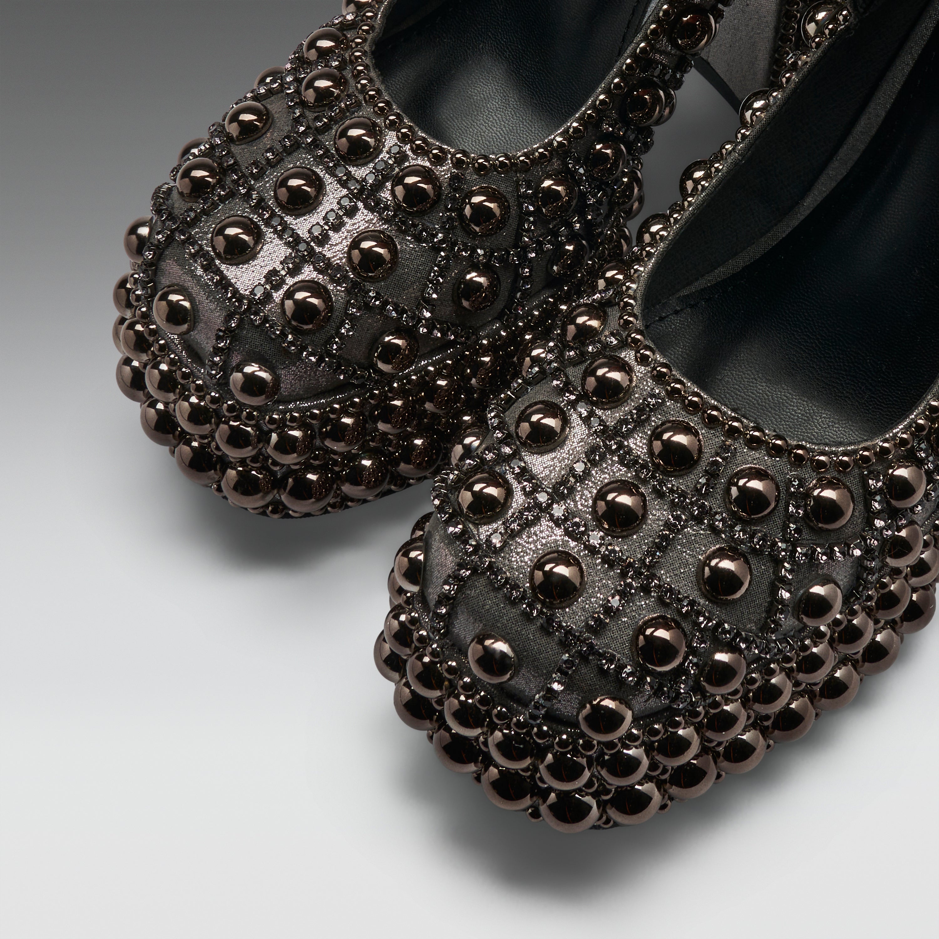 A pair of black leather ballet flats heavily embellished with metallic bronze and dark brown beaded studs, arranged on a light gray studio background.