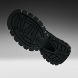 A close-up studio shot of a black rubber shoe sole, showing the tread pattern and embossed branding, set against a dark gray gradient background.