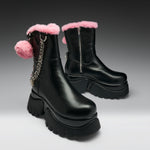 A pair of black leather ankle boots with pink faux-fur trim, chunky black platform soles, and a pink pom-pom charm with a silver chain on the side, set against a dark gray gradient background.