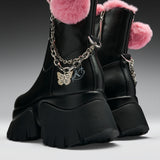 Black leather ankle boots with chunky lug soles, pink faux-fur trim, silver chain details, and a gold butterfly charm, shown on a dark gray studio background.