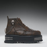 A dark brown textured leather ankle boot with a chunky black platform sole, decorated with silver skull hardware, a hanging chain, and metal studs, shown against a dark gray gradient background.