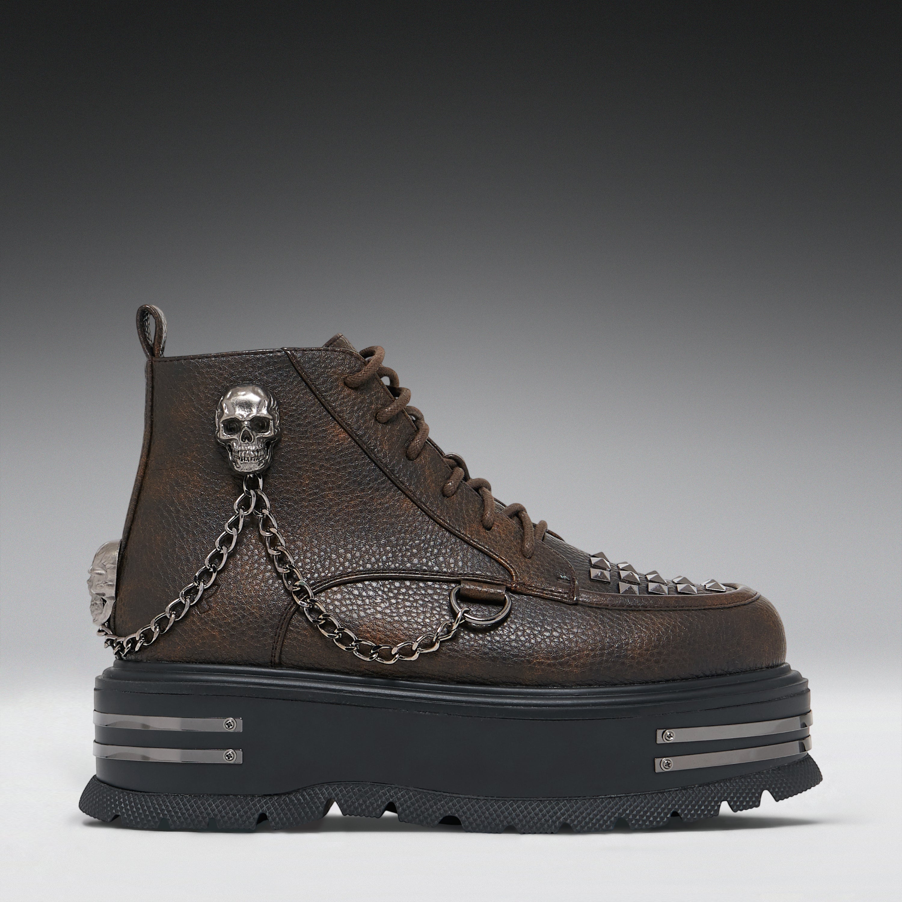 A dark brown textured leather ankle boot with a chunky black platform sole, decorated with silver skull hardware, a hanging chain, and metal studs, shown against a dark gray gradient background.