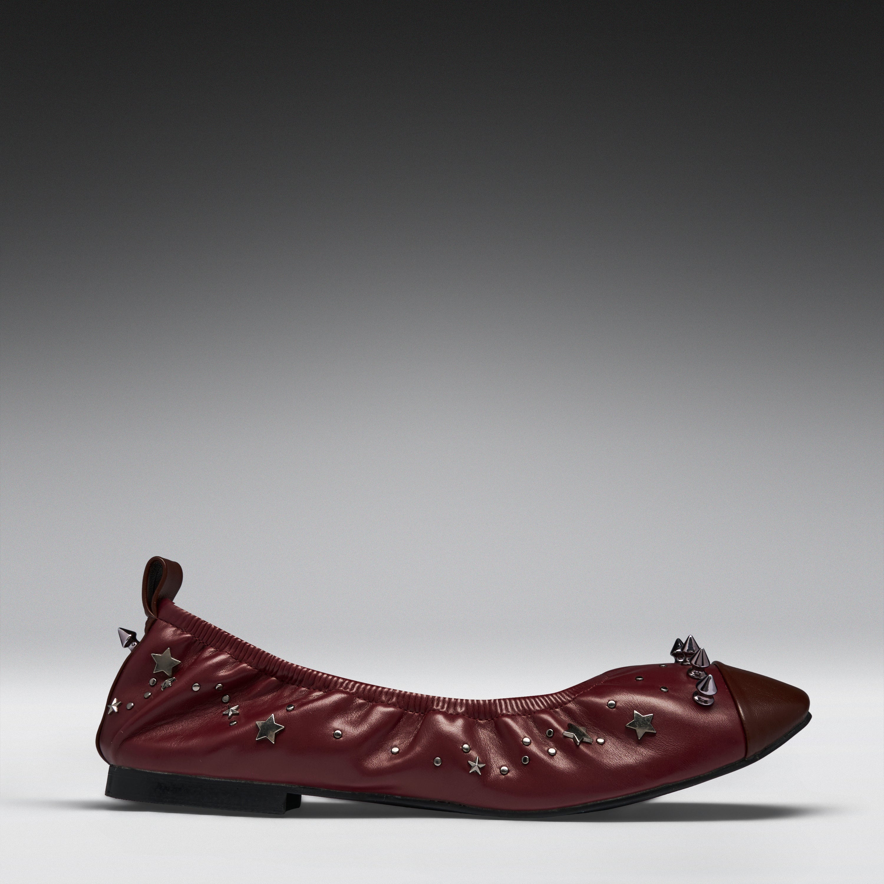 A single burgundy leather ballet flat with star-shaped metallic embellishments and a dark brown pointed toe, shown in profile against a gray gradient studio background.