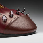 A close-up of a burgundy leather shoe featuring metallic spiked studs on the toe area, set against a dark gray gradient background.