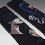 A pair of black knit socks featuring printed anime-style character illustrations, shown diagonally on a light gray background.