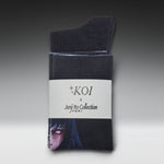 A pair of dark charcoal gray crew socks folded and wrapped in a white paper band. The band features the text 'KOI x Junji Tko Collection' and a printed illustration of a character from the anime Junji No Naka de Amu.