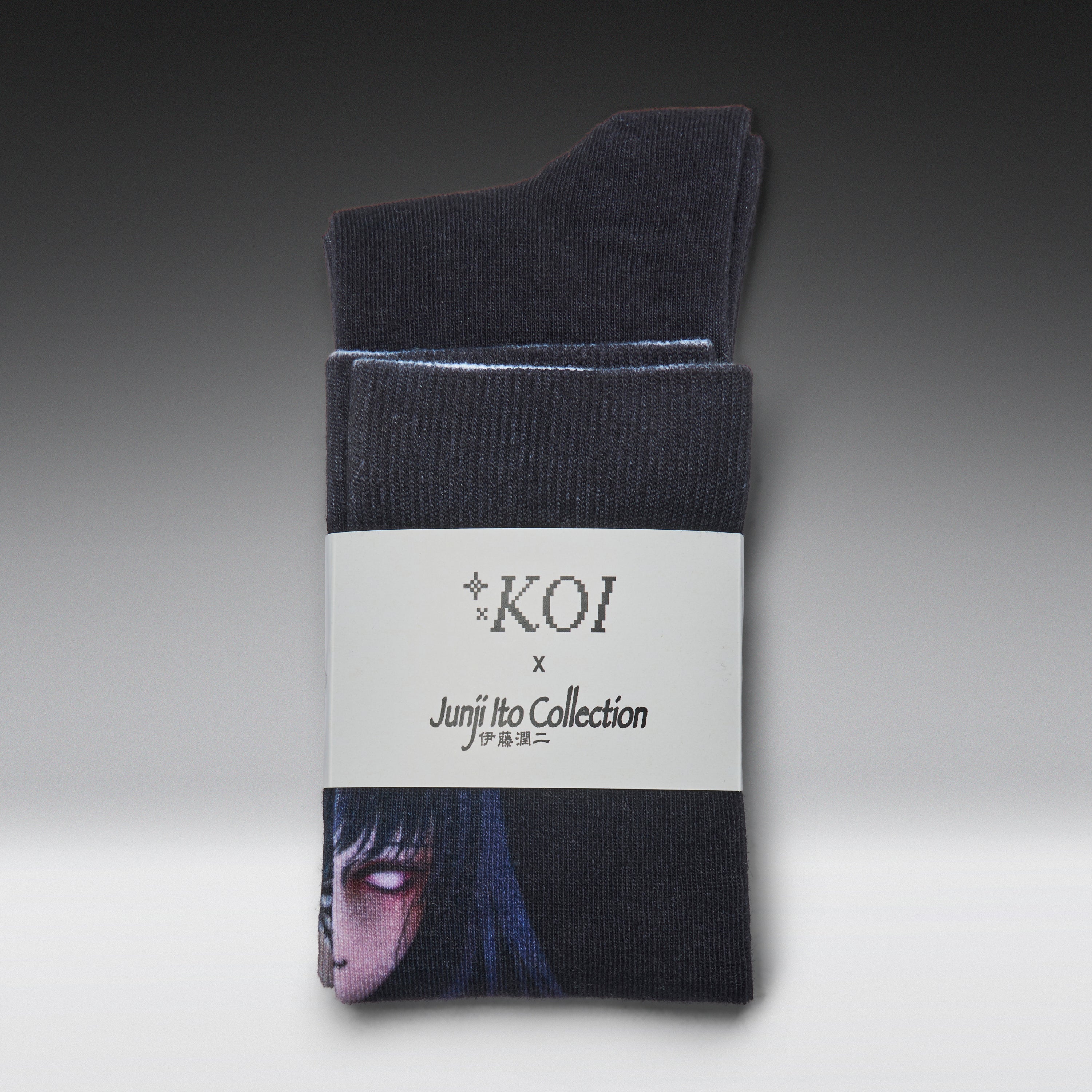 A pair of dark charcoal gray crew socks folded and wrapped in a white paper band. The band features the text 'KOI x Junji Tko Collection' and a printed illustration of a character from the anime Junji No Naka de Amu.