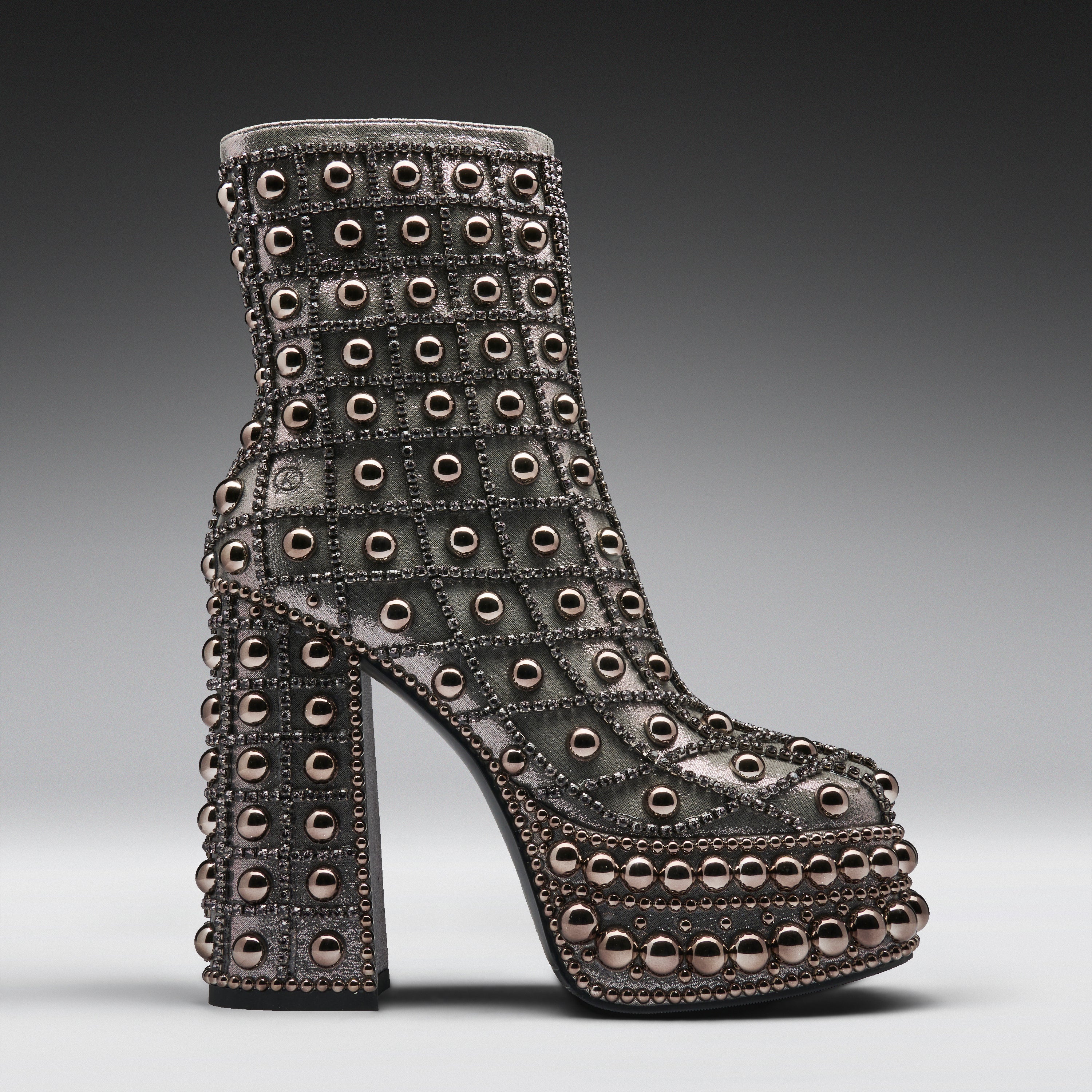A single high-heeled platform ankle boot covered in rows of metallic studs and rhinestones, shown in profile against a dark gray gradient studio background.