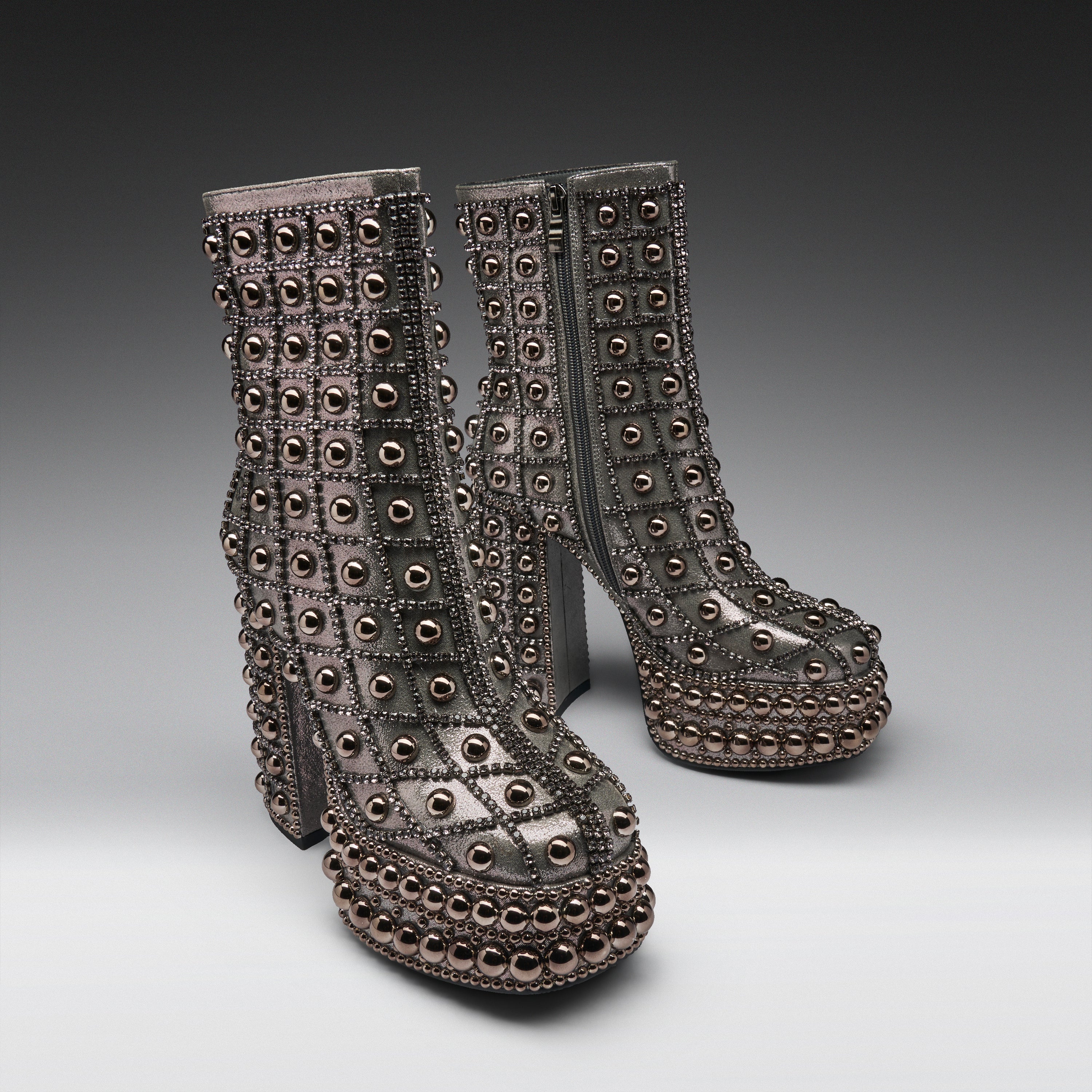 A pair of metallic silver platform ankle boots heavily embellished with rows of round dark metallic studs and rhinestones, set against a dark-to-light gray studio background.