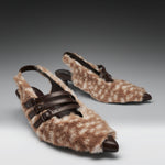 A pair of pointed-toe slingback kitten heels covered in brown and beige spotted faux fur, with dark brown leather straps and buckle details, photographed on a dark-to-light gray gradient studio background.