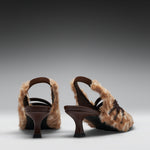 A pair of women's slingback high-heeled sandals featuring brown straps and thick, fluffy beige faux-fur uppers, shown from a rear three-quarter angle on a dark-to-light gray gradient studio background.