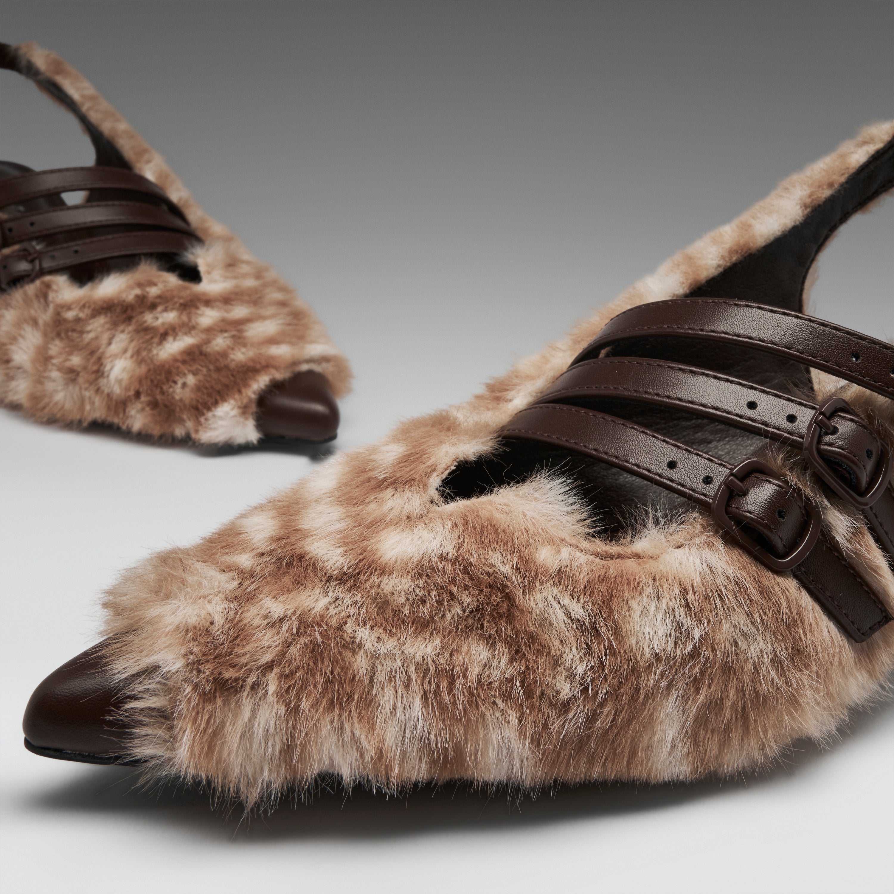 A close-up of a pair of pointed-toe mule flats covered in brown and beige mottled faux fur, with dark brown leather straps and buckle details, on a neutral gray background.